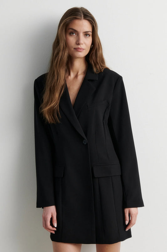 A woman modeling a black double-breasted blazer-style mini dress with a wrap front and flap pockets, photographed against a plain light gray studio background.