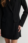 A close-up of a model wearing a black tailored blazer-style mini dress with structured shoulders, a single front button, flap pockets, and a straight skirt, against a light neutral background.