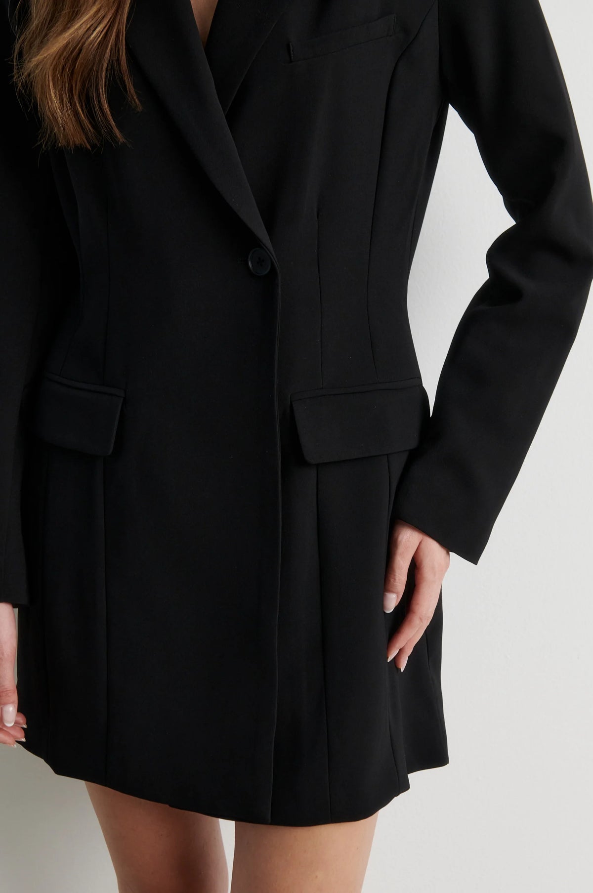 A close-up of a model wearing a black tailored blazer-style mini dress with structured shoulders, a single front button, flap pockets, and a straight skirt, against a light neutral background.