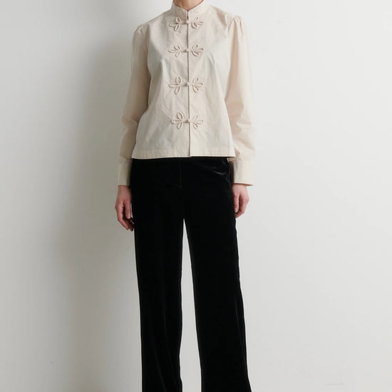 A model stands against a plain white wall wearing a cream-colored long-sleeve jacket with decorative buttons and black wide-leg trousers.