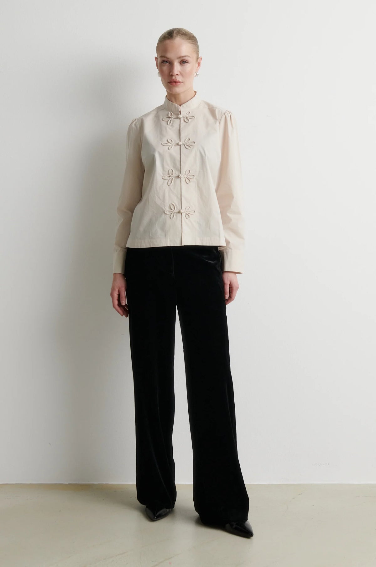 A model stands against a plain white wall wearing a cream-colored long-sleeve jacket with decorative buttons and black wide-leg trousers.