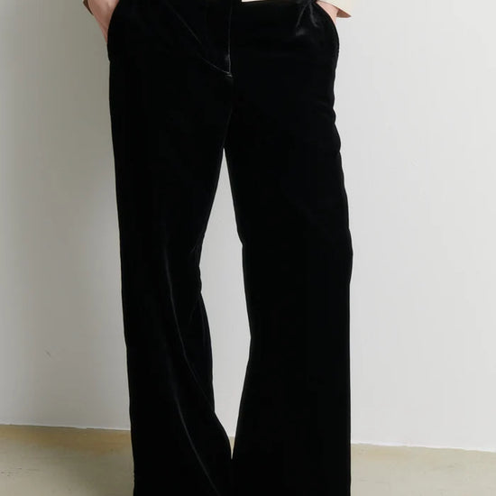 A person wearing black wide-leg velvet pants with a cream-colored jacket, standing against a plain off-white wall.