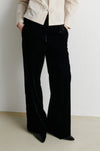 A person wearing black wide-leg velvet pants with a cream-colored jacket, standing against a plain off-white wall.