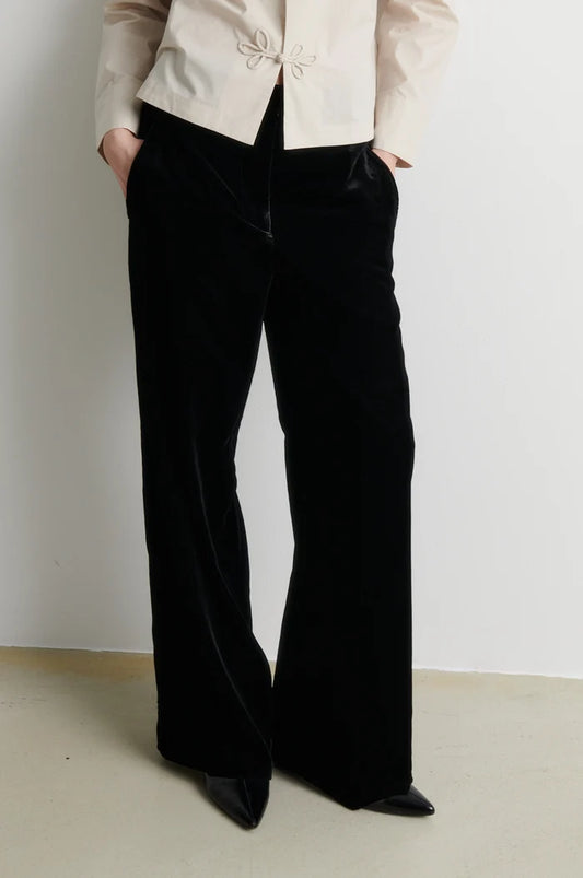 A person wearing black wide-leg velvet pants with a cream-colored jacket, standing against a plain off-white wall.