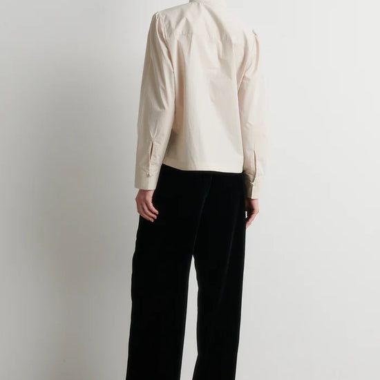 A model stands against a plain white wall wearing a cream-colored long-sleeve jacket with decorative buttons and black wide-leg trousers.