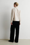 A model stands against a plain white wall wearing a cream-colored long-sleeve jacket with decorative buttons and black wide-leg trousers.