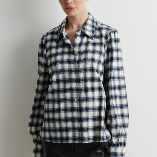 A woman modeling a long-sleeve button-front plaid shirt in navy, white, and light gray, paired with black pants, against a plain light gray studio background.
