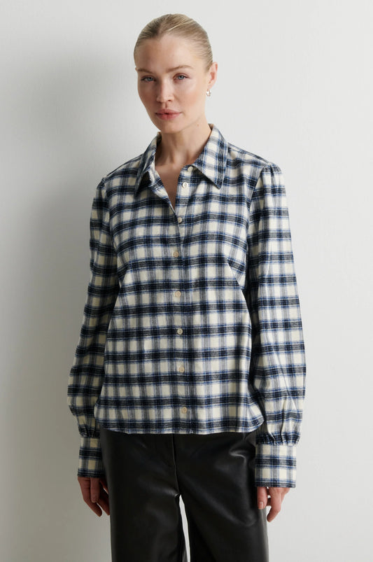 A woman modeling a long-sleeve button-front plaid shirt in navy, white, and light gray, paired with black pants, against a plain light gray studio background.