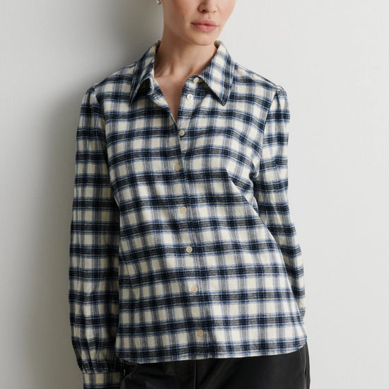 A woman wearing a long-sleeve button-up plaid shirt in navy, white, and black, paired with black pants, standing against a plain light gray wall.
