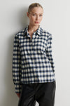 A woman wearing a long-sleeve button-up plaid shirt in navy, white, and black, paired with black pants, standing against a plain light gray wall.
