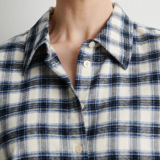 A woman wearing a long-sleeve button-up plaid shirt in navy, white, and black, paired with black pants, standing against a plain light gray wall.