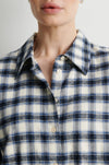 A woman wearing a long-sleeve button-up plaid shirt in navy, white, and black, paired with black pants, standing against a plain light gray wall.