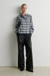 A woman wearing a long-sleeve button-up plaid shirt in navy, white, and black, paired with black pants, standing against a plain light gray wall.