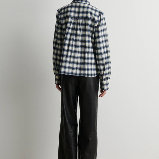A model viewed from the back wearing a navy and white gingham plaid button-up shirt and black wide-leg trousers, standing against a plain light gray wall.