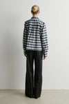 A model viewed from the back wearing a navy and white gingham plaid button-up shirt and black wide-leg trousers, standing against a plain light gray wall.
