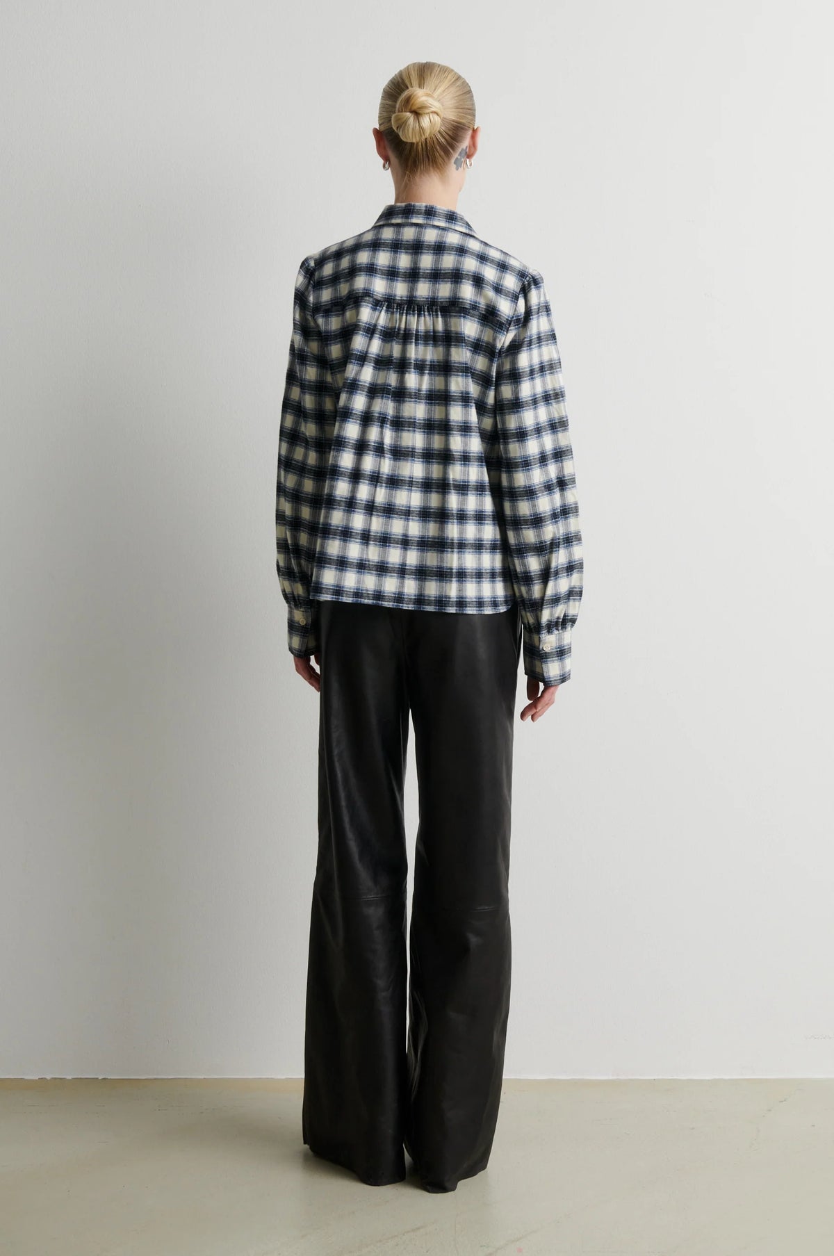 A model viewed from the back wearing a navy and white gingham plaid button-up shirt and black wide-leg trousers, standing against a plain light gray wall.