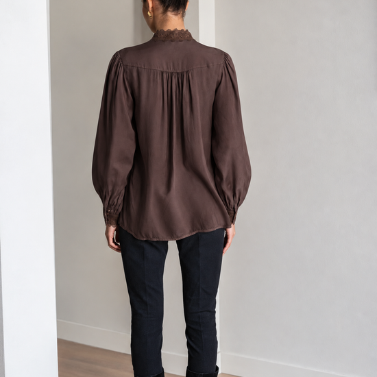 A woman viewed from behind wearing a dark brown long-sleeve blouse with gathered detailing, paired with black pants and black knee-high boots, standing in a minimalist room with light gray walls and a wood floor.