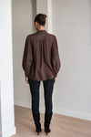 A woman viewed from behind wearing a dark brown long-sleeve blouse with gathered detailing, paired with black pants and black knee-high boots, standing in a minimalist room with light gray walls and a wood floor.
