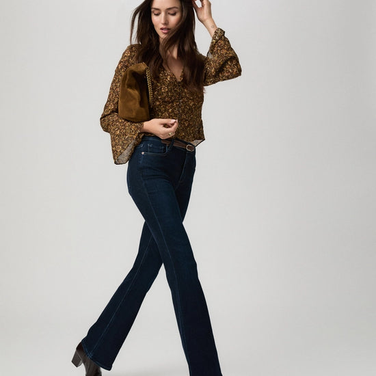 A woman modeling a brown and gold patterned blouse with bell sleeves, dark blue jeans, and dark brown heeled ankle boots, carrying a brown bag against a light gray studio background.