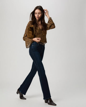A woman modeling a brown and gold patterned blouse with bell sleeves, dark blue jeans, and dark brown heeled ankle boots, carrying a brown bag against a light gray studio background.