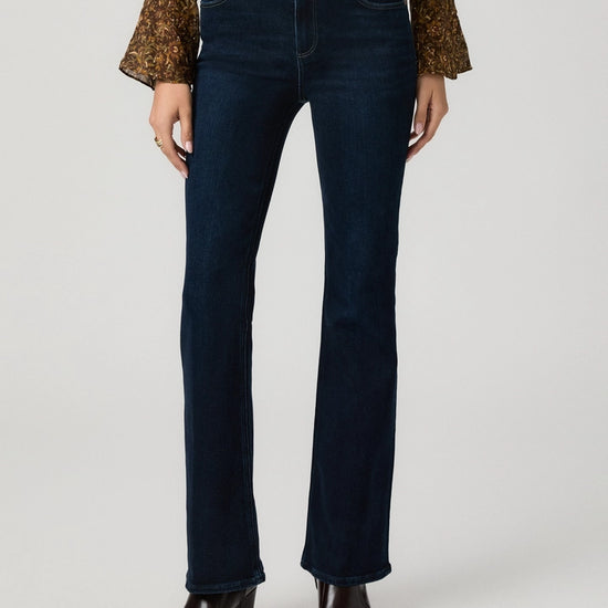 A model wearing dark blue straight-leg denim jeans, a brown patterned long-sleeve top, and dark brown leather ankle boots, shown from the waist down on a light gray studio background.