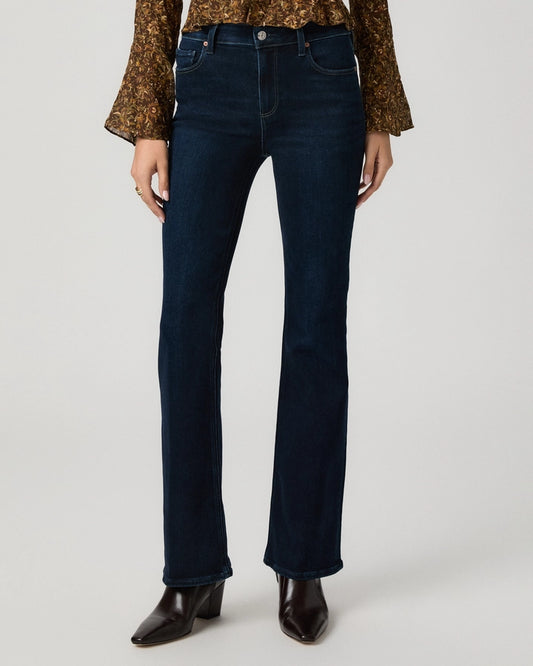 A model wearing dark blue straight-leg denim jeans, a brown patterned long-sleeve top, and dark brown leather ankle boots, shown from the waist down on a light gray studio background.