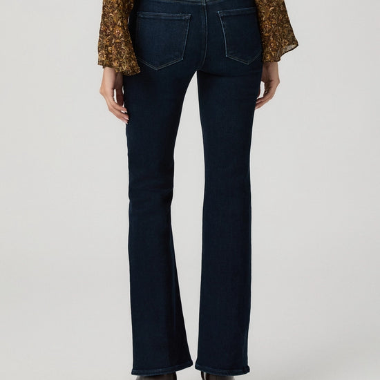 Rear view of a model wearing dark blue flare jeans, a brown patterned long-sleeve top, and dark brown heeled shoes against a light gray studio background.