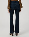 Rear view of a model wearing dark blue flare jeans, a brown patterned long-sleeve top, and dark brown heeled shoes against a light gray studio background.