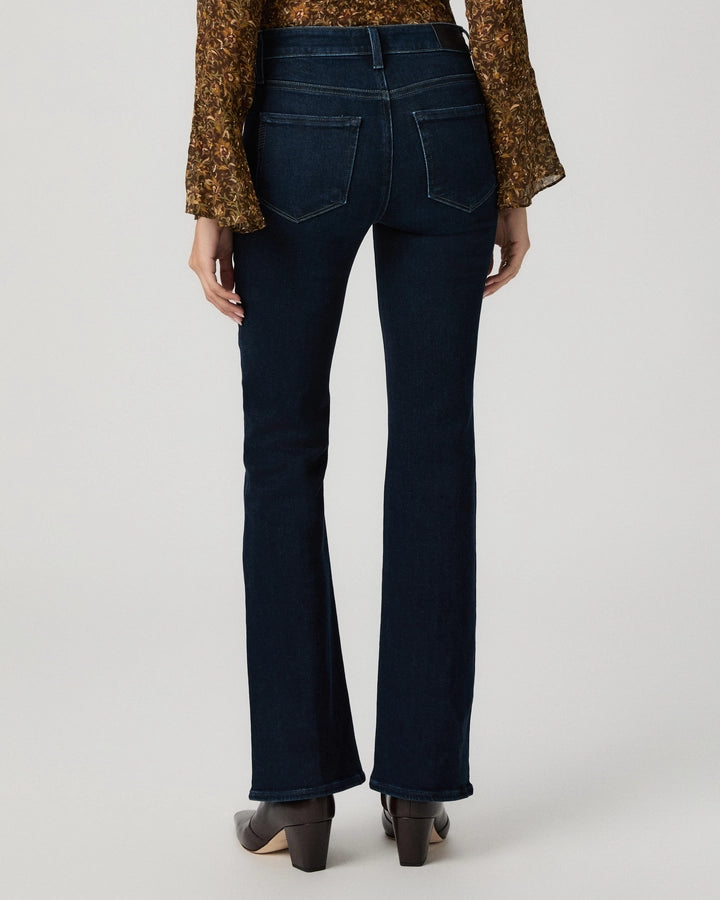 Rear view of a model wearing dark blue flare jeans, a brown patterned long-sleeve top, and dark brown heeled shoes against a light gray studio background.