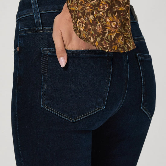 Close-up back view of a person wearing dark indigo denim jeans with a floral-patterned blouse tucked into the waistband, against a light gray studio background.