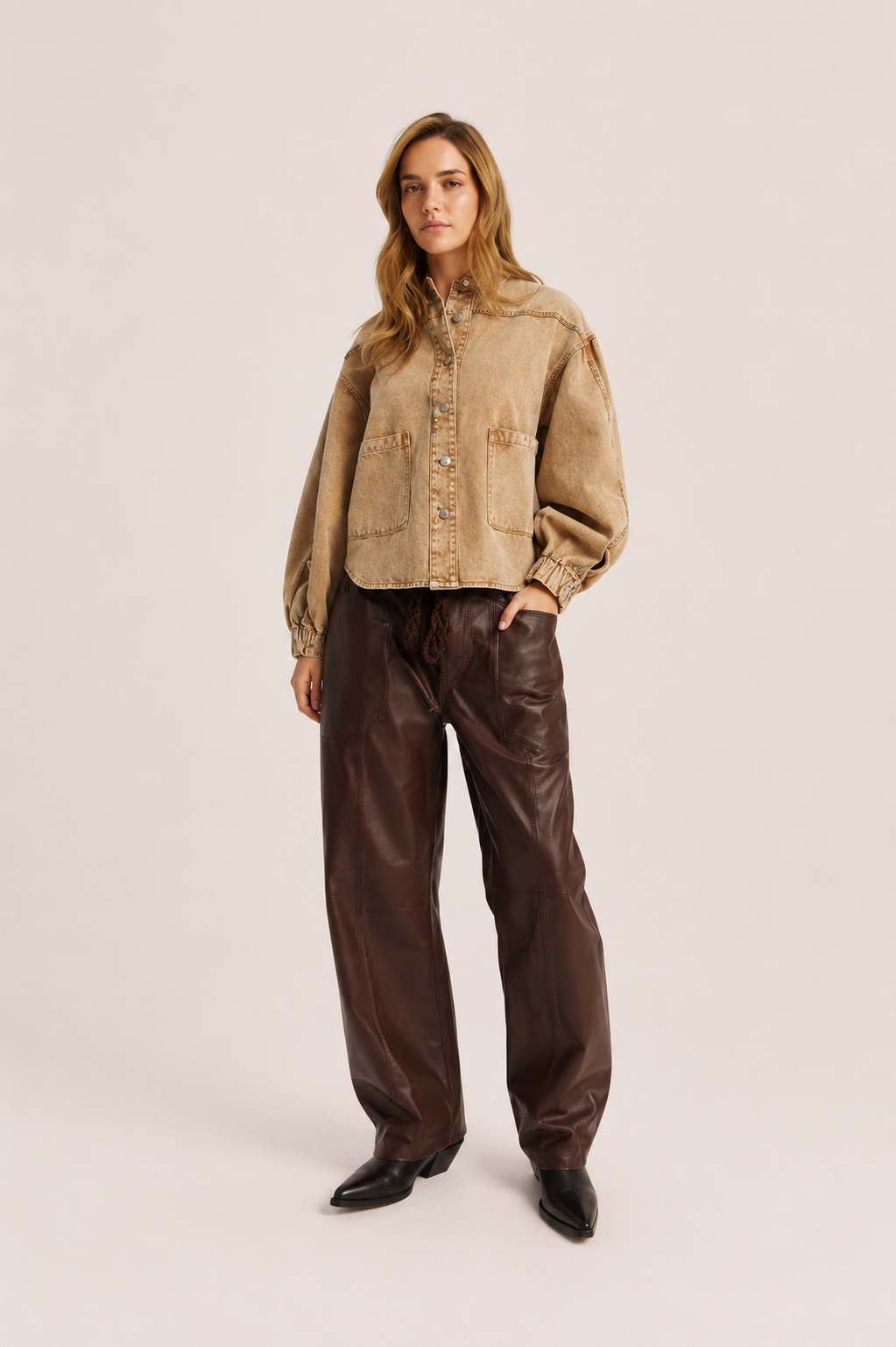 A woman modeling a cropped tan suede-like button-up jacket with voluminous sleeves, paired with dark brown wide-leg faux-leather pants and black pointed boots, against a plain light beige studio background.