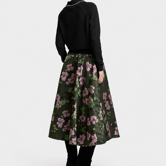 A model is shown from the back wearing a black knit sweater with a pearl collar and a dark green midi skirt featuring a pink and purple floral pattern. She is wearing black ankle boots against a plain light gray studio background.