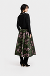 A model is shown from the back wearing a black knit sweater with a pearl collar and a dark green midi skirt featuring a pink and purple floral pattern. She is wearing black ankle boots against a plain light gray studio background.