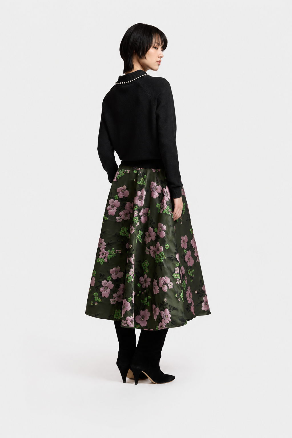 A model is shown from the back wearing a black knit sweater with a pearl collar and a dark green midi skirt featuring a pink and purple floral pattern. She is wearing black ankle boots against a plain light gray studio background.