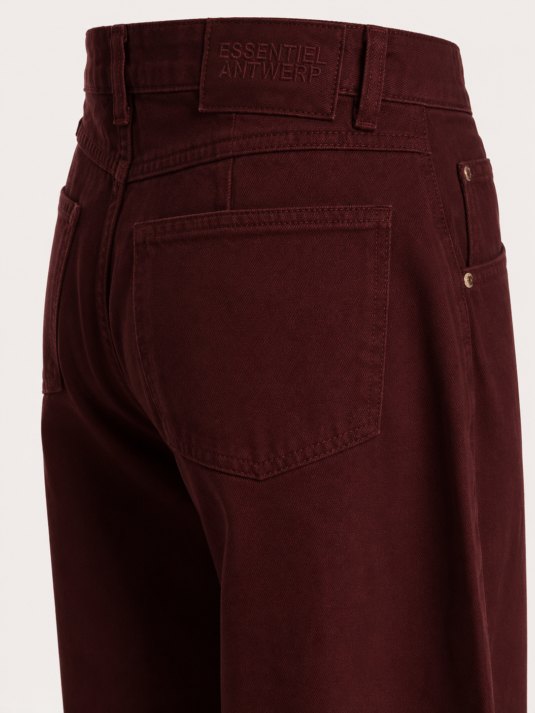 Close-up of the back waistband and rear pocket area of dark burgundy denim jeans, showing the brand label 'ESSENTIEL ANTWERP' and a button closure.