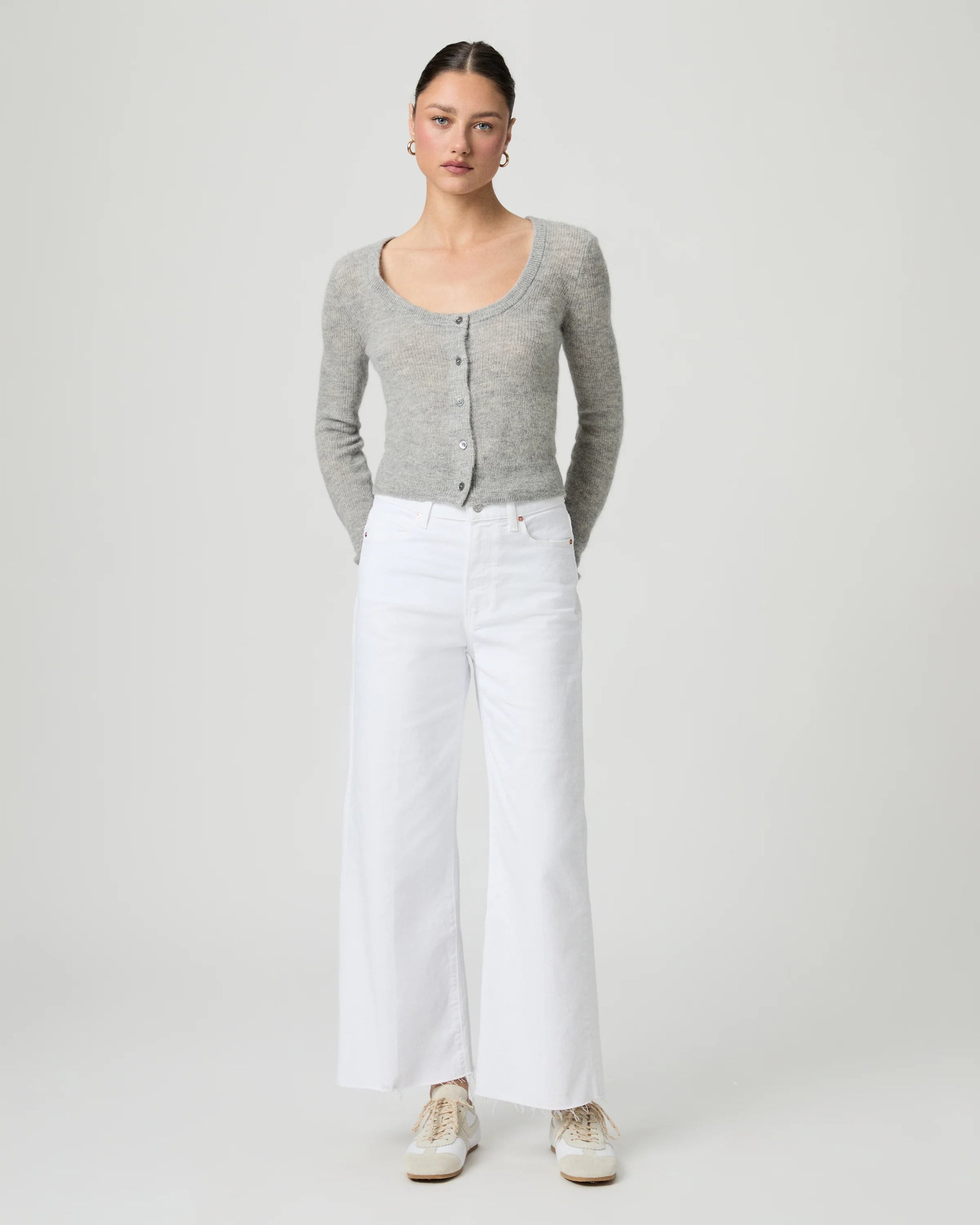 Woman wearing a gray cardigan and white paige jeans on a plain background
