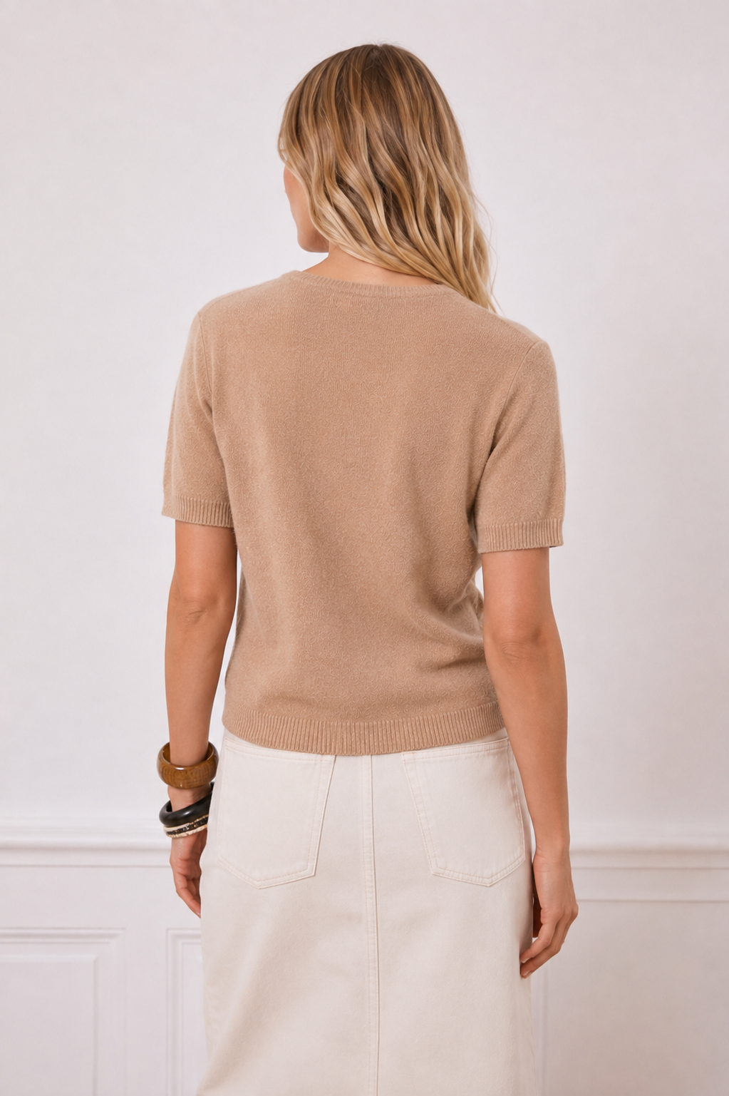 A woman shown from the back wearing a short-sleeve knit sweater in a light tan/beige color, paired with an off-white skirt, against a white wall with decorative molding.