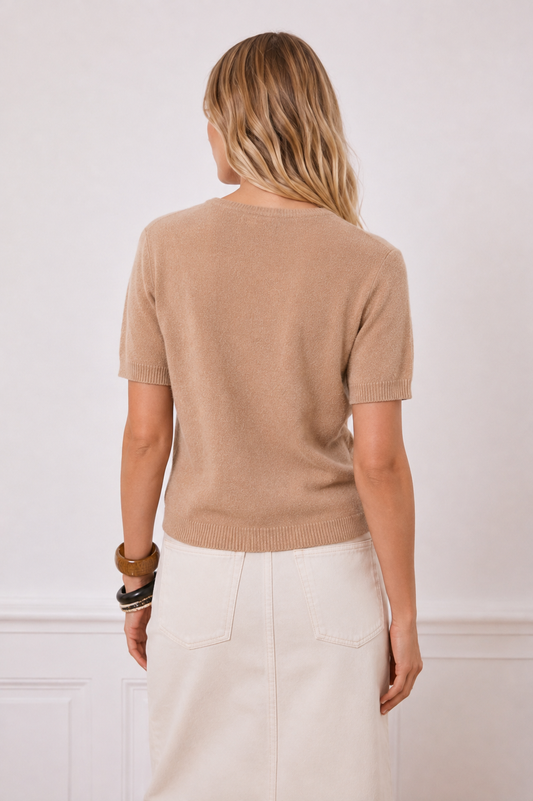 A woman shown from the back wearing a short-sleeve knit sweater in a light tan/beige color, paired with an off-white skirt, against a white wall with decorative molding.