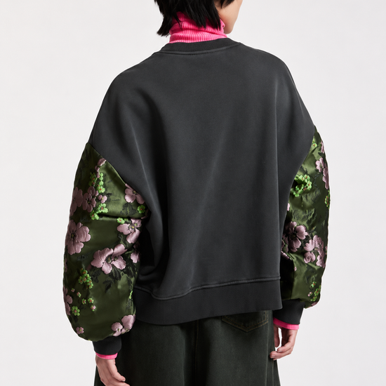A model shown from the back wearing a black sweatshirt with green floral patterned balloon sleeves, layered over a bright pink turtleneck and dark wide-leg trousers, against a plain light gray background.