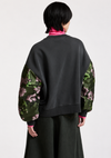 A model shown from the back wearing a black sweatshirt with green floral patterned balloon sleeves, layered over a bright pink turtleneck and dark wide-leg trousers, against a plain light gray background.