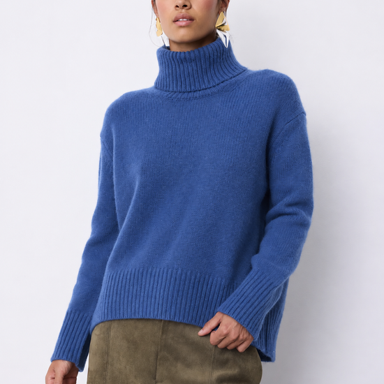 A woman wearing a blue ribbed turtleneck sweater and olive green trousers, standing against a plain light gray studio background.