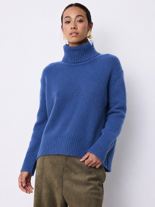 A woman wearing a blue ribbed turtleneck sweater and olive green trousers, standing against a plain light gray studio background.