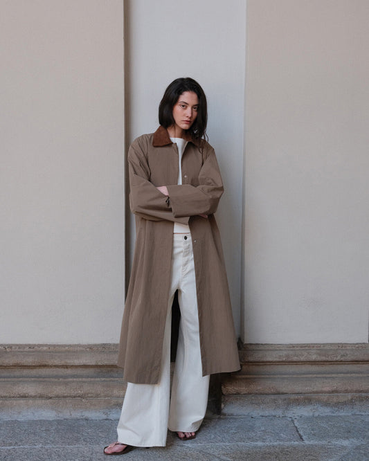 A woman stands outdoors against a beige stone wall, wearing a long taupe-brown trench coat with a brown collar, white wide-leg trousers, and pink sandals.
