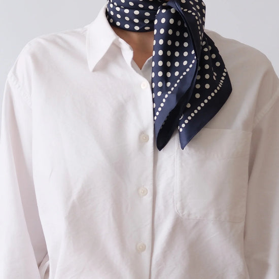 Person wearing a navy blue scarf with white polka dots over a white shirt against a plain background