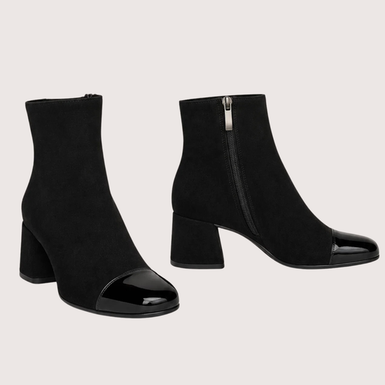 A pair of black ankle boots with a block heel and glossy patent leather toe caps, shown against a solid light beige background.
