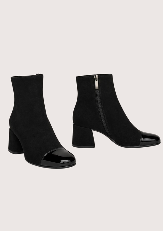 A pair of black ankle boots with a block heel and glossy patent leather toe caps, shown against a solid light beige background.