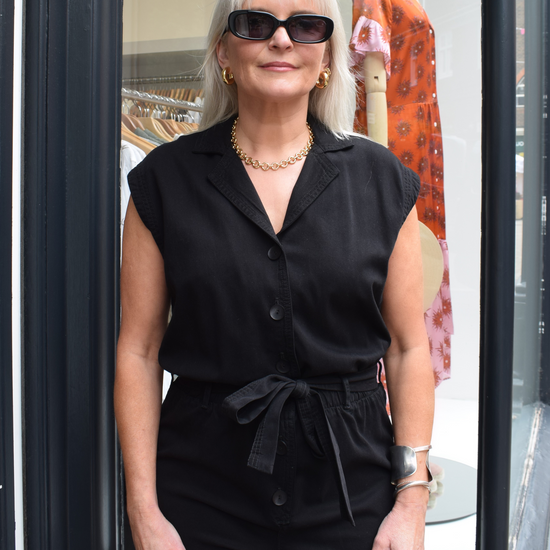 A photo of a black, sleevless playsuit with a tie belt and gold accesiores