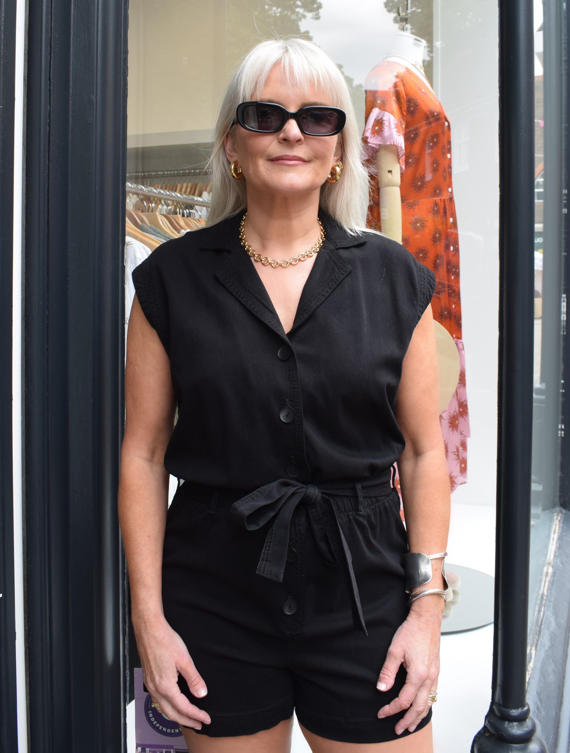 A photo of a black, sleevless playsuit with a tie belt and gold accesiores