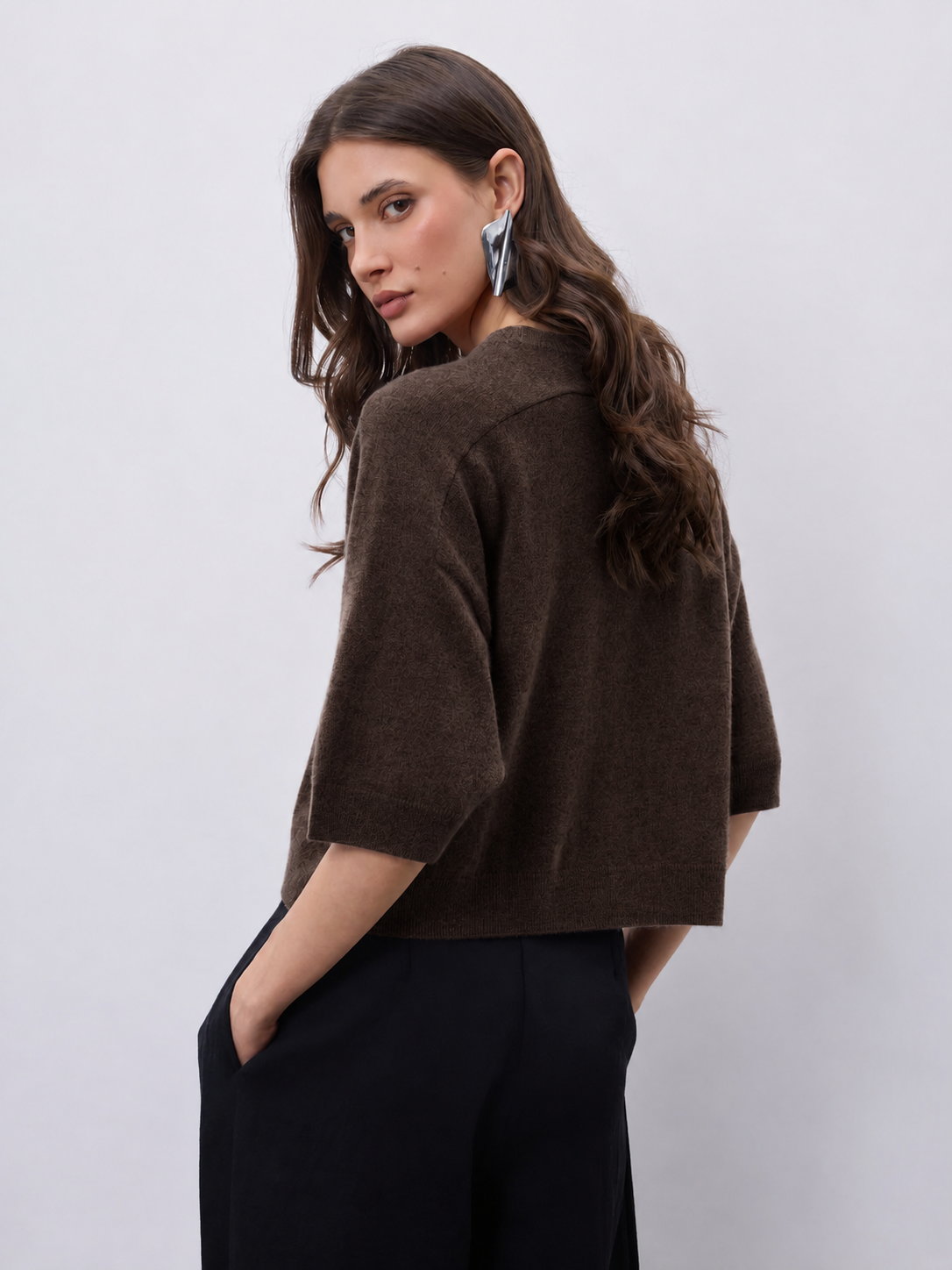 model wearing a dark brown short sleeve knit, with black trousers on a plain background 