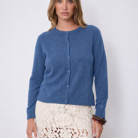 A woman modeling a blue long-sleeve button-front cardigan sweater paired with a cream lace skirt, standing against a plain light gray studio background.
